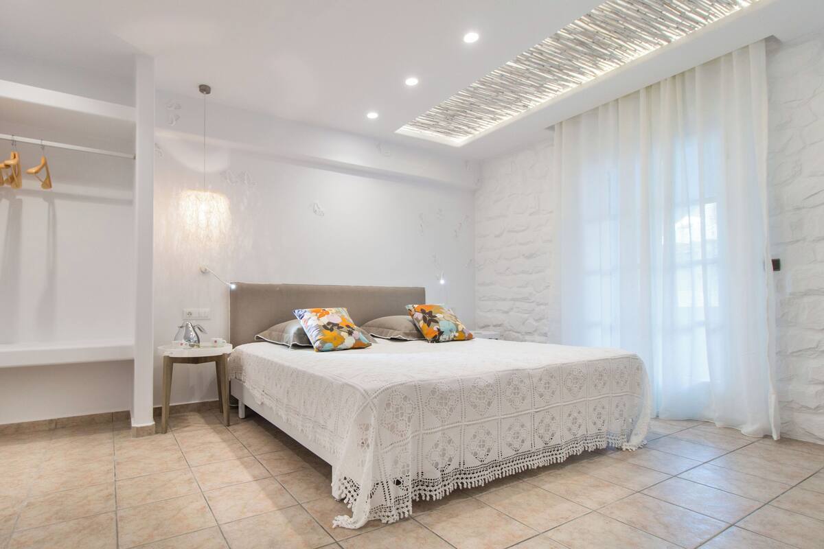Une chambre moderne avec un grand lit, une table de chevet, une étagère murale avec des vêtements et une fenêtre ornée de rideaux légers.