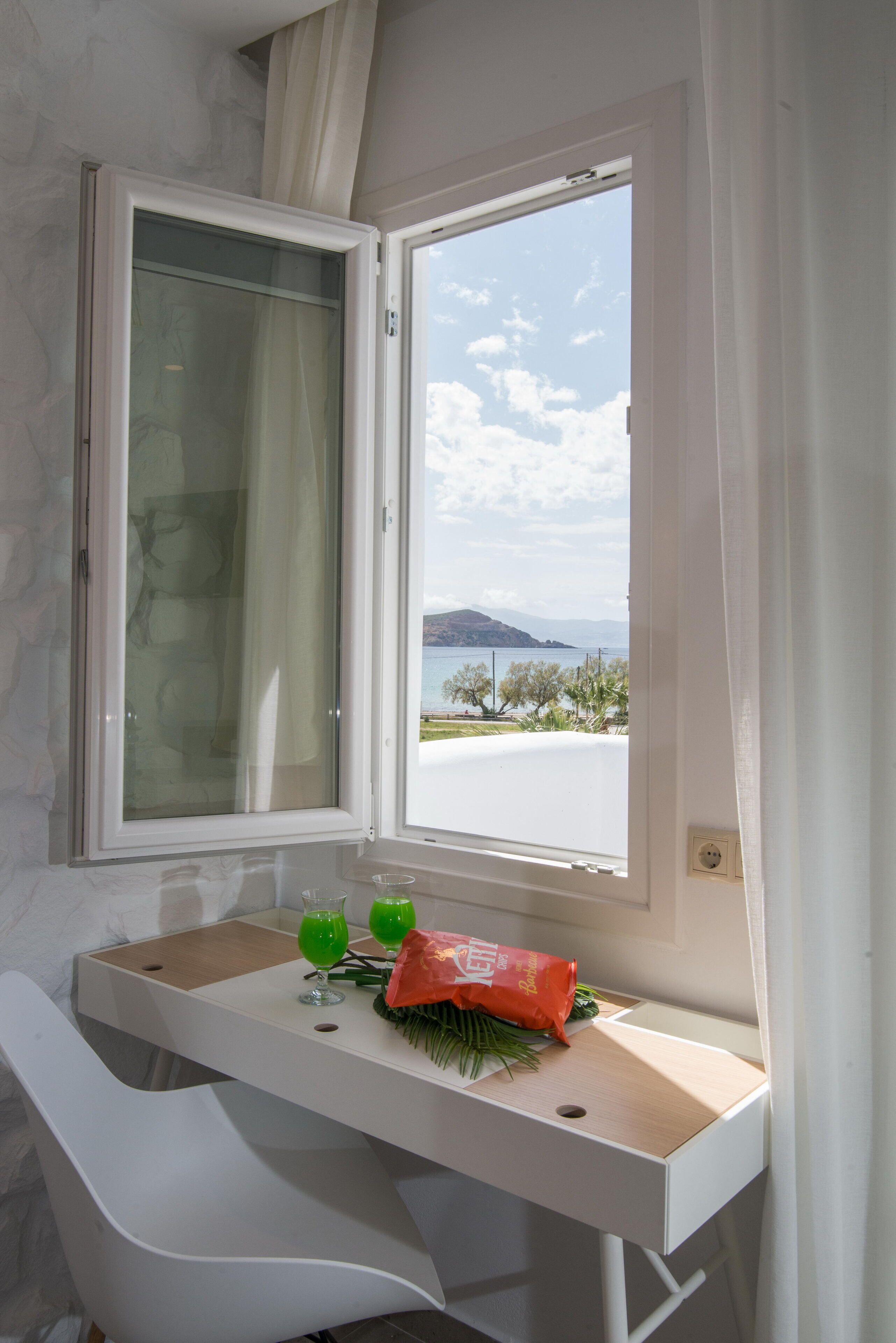 Una habitación con escritorio, silla, ventana que ofrece vistas a un cuerpo de agua y montañas, y una bolsa de bocadillos sobre el escritorio.