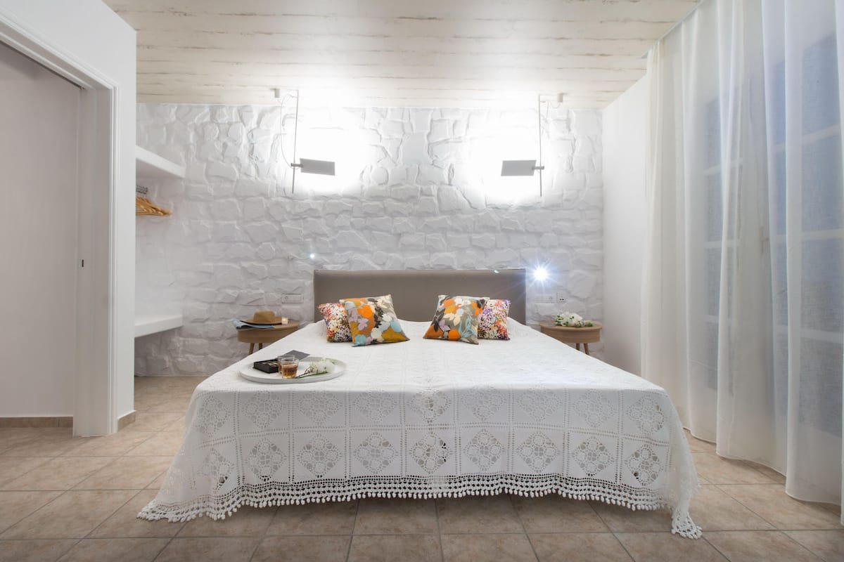 Une chambre avec un mur en pierre, un lit recouvert d’une courtepointe blanche, deux tables de chevet et une fenêtre avec des rideaux.