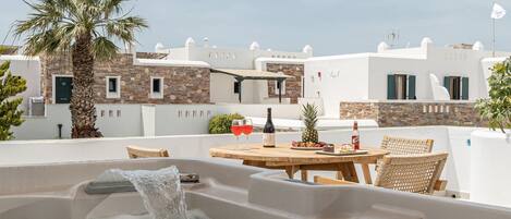 A rooftop hot tub with a wooden pergola overhead, surrounded by white buildings and a palm tree.