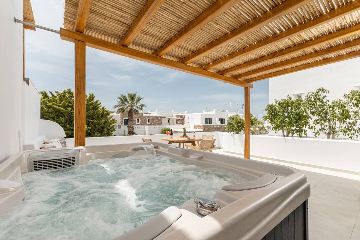 Un bain à remous sous une pergola en bois, avec vue sur un bâtiment blanc et une végétation luxuriante en arrière-plan.