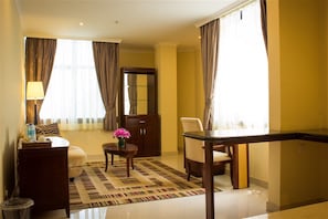 A hotel room with a sofa, a small table with a vase of flowers, a desk, and a chair. There is a large mirror on the wall and a window with curtains.