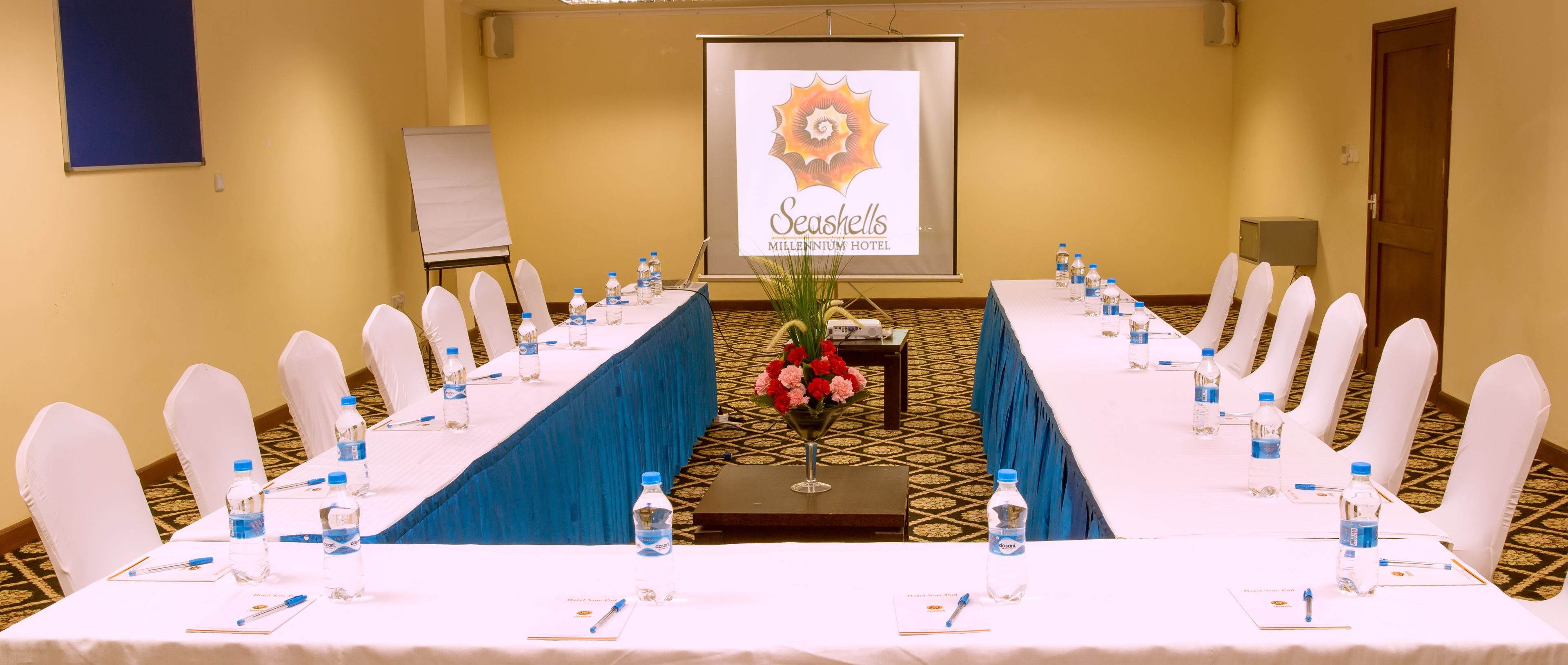 A conference room at the Seashells Millennium Hotel with a long table, white chairs, water bottles, and a projector screen in the background.