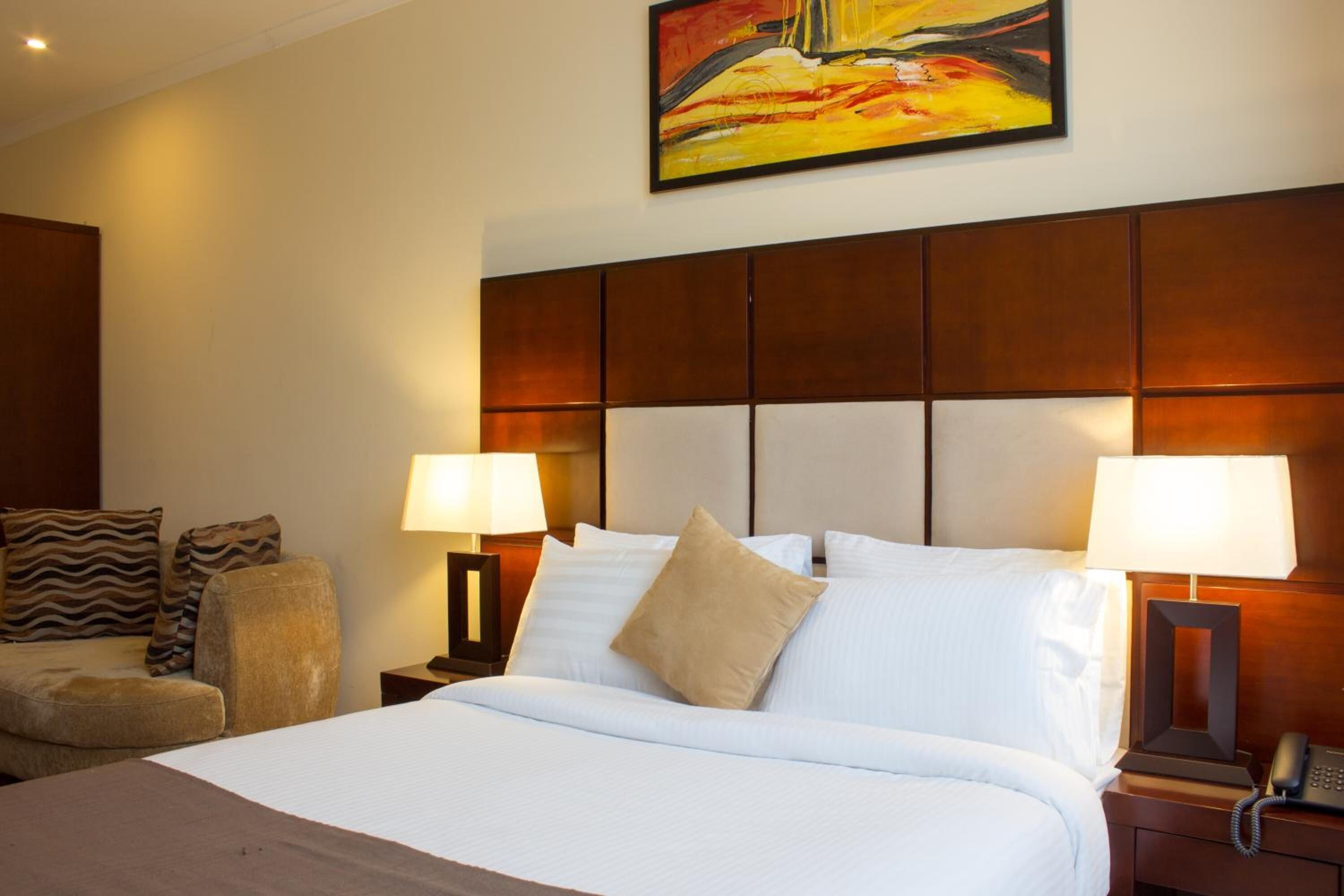 A hotel room with a large bed, two bedside tables with lamps, a headboard, and a painting on the wall.