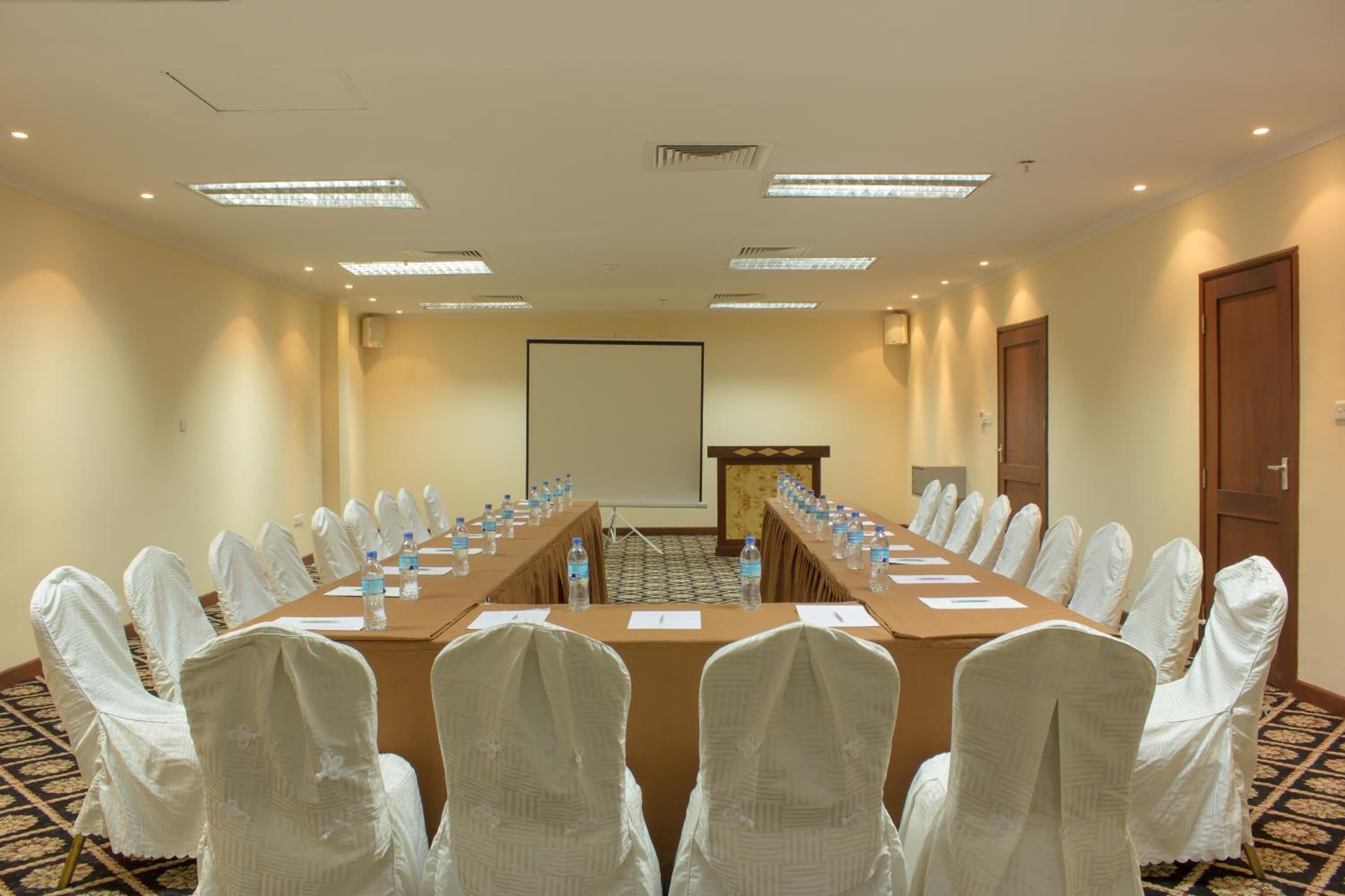 Un salón de conferencias con una larga mesa, sillas, una pantalla de proyector y un atril.