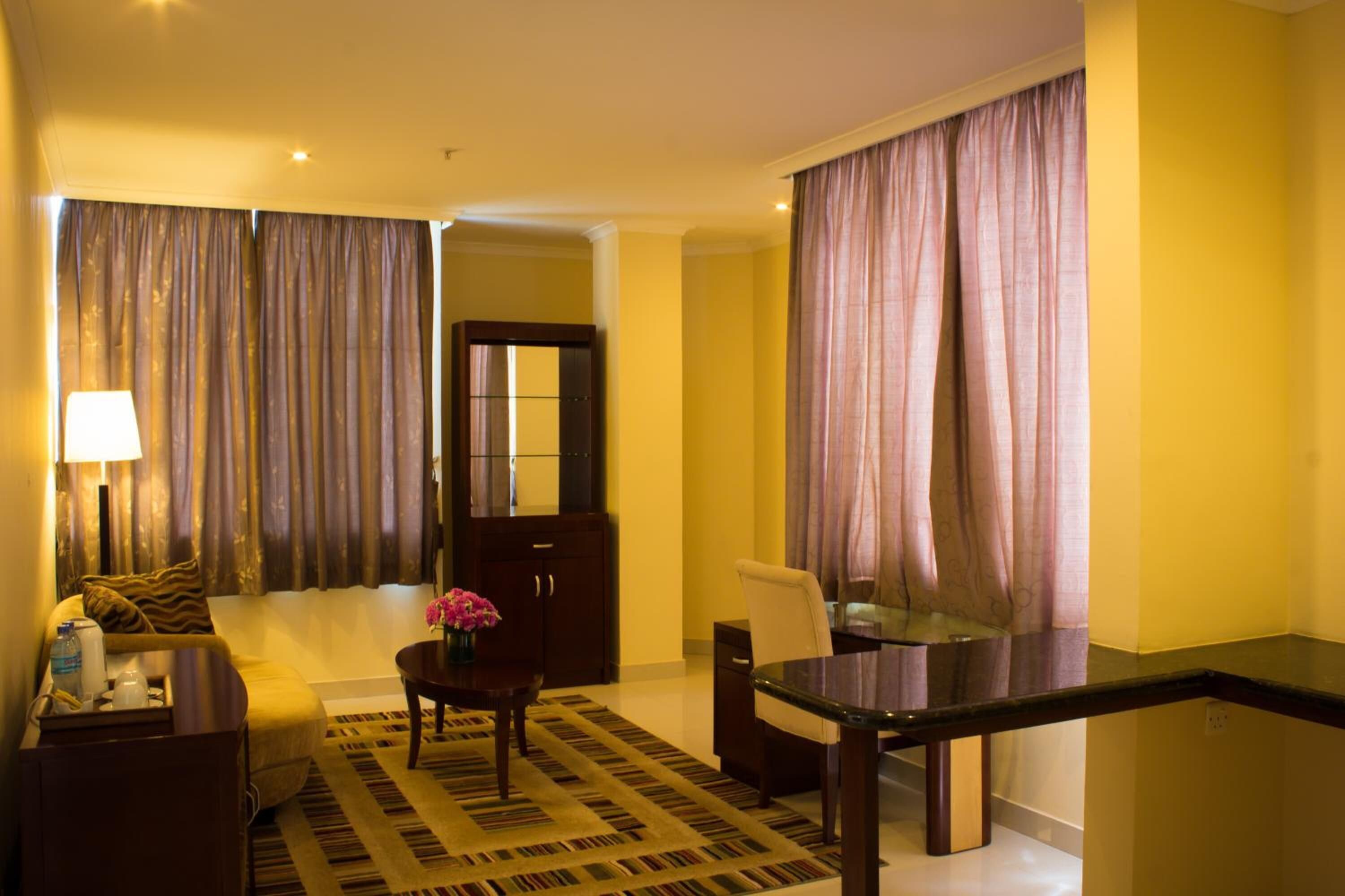 A hotel room with a sofa, a dining table, a coffee table, and a lamp. There is a mirror on the wall and a door leading to another room.