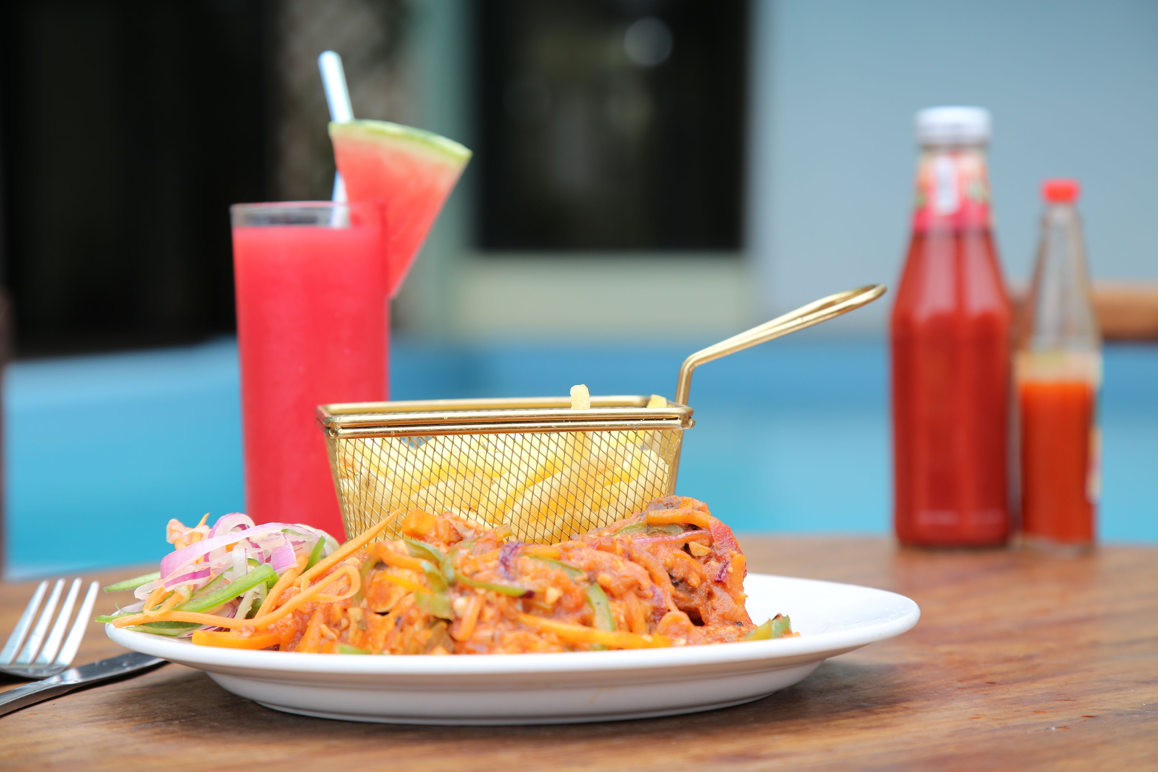 Un piatto di noodles con verdure, un bicchiere da cocktail e due bottiglie di salsa su un tavolo di legno.