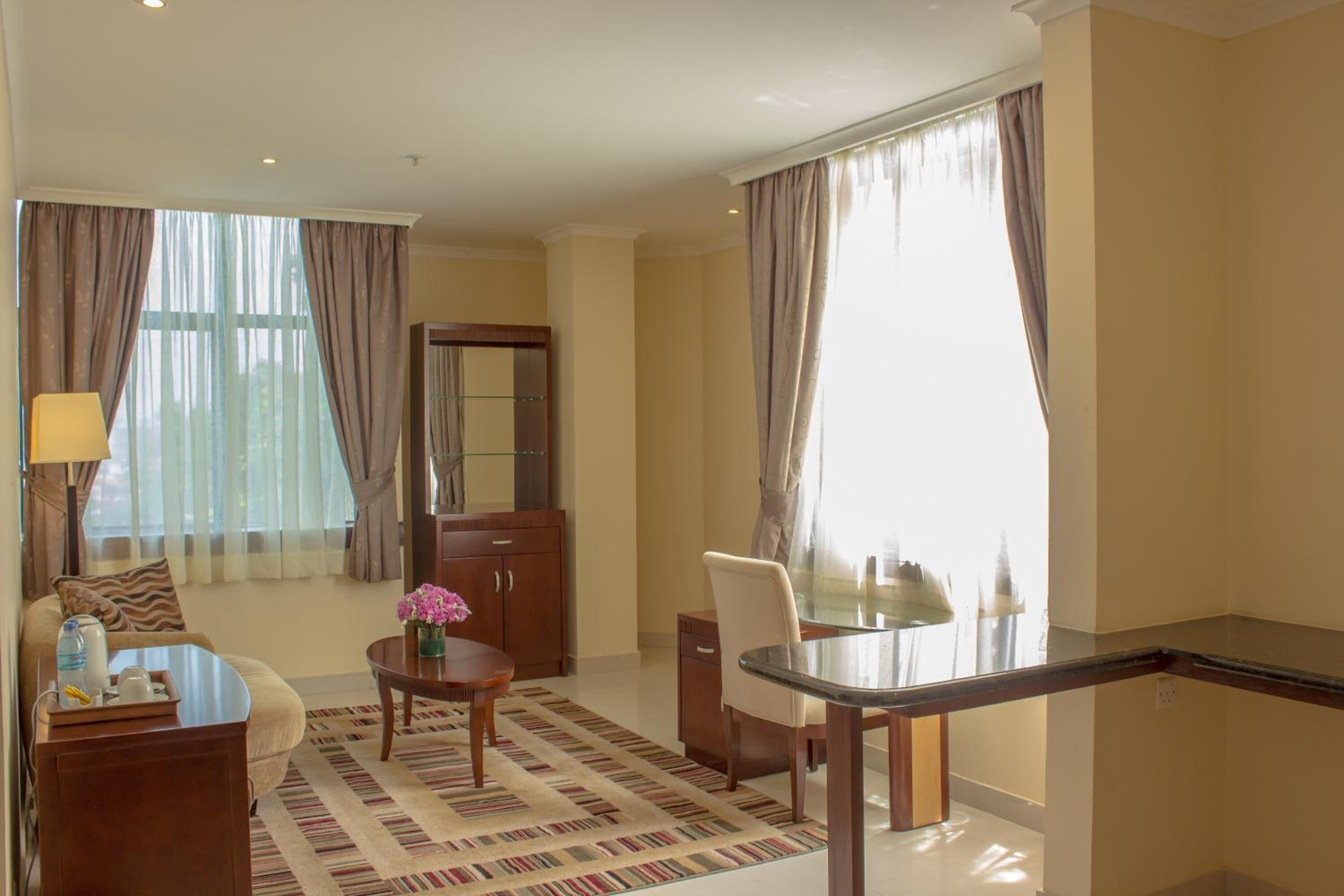 Una habitación de hotel con sofá, una mesa de comedor, una alfombra pequeña, un armario y un ventanal con cortinas.