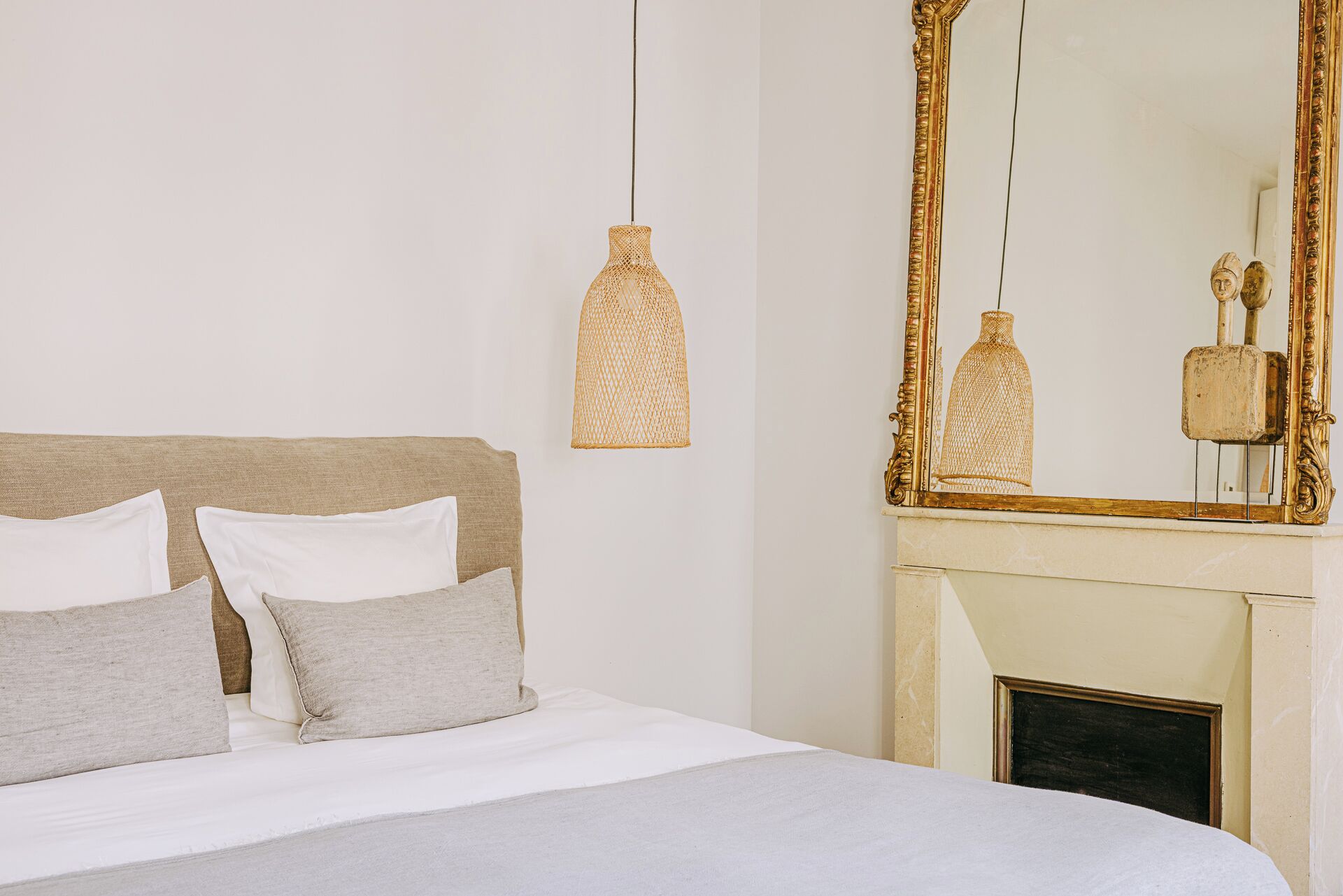 Un dormitorio con cama, almohadas, una lámpara colgante y un espejo grande sobre una chimenea.