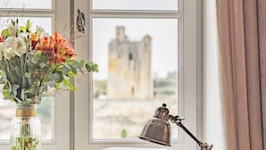 Una habitación con un ventanal grande, un escritorio con libros, una lámpara, un florero con flores y vistas a un castillo.