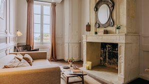 Una habitación con chimenea, un sofá, una mesa con libros y una ventana con cortinas.