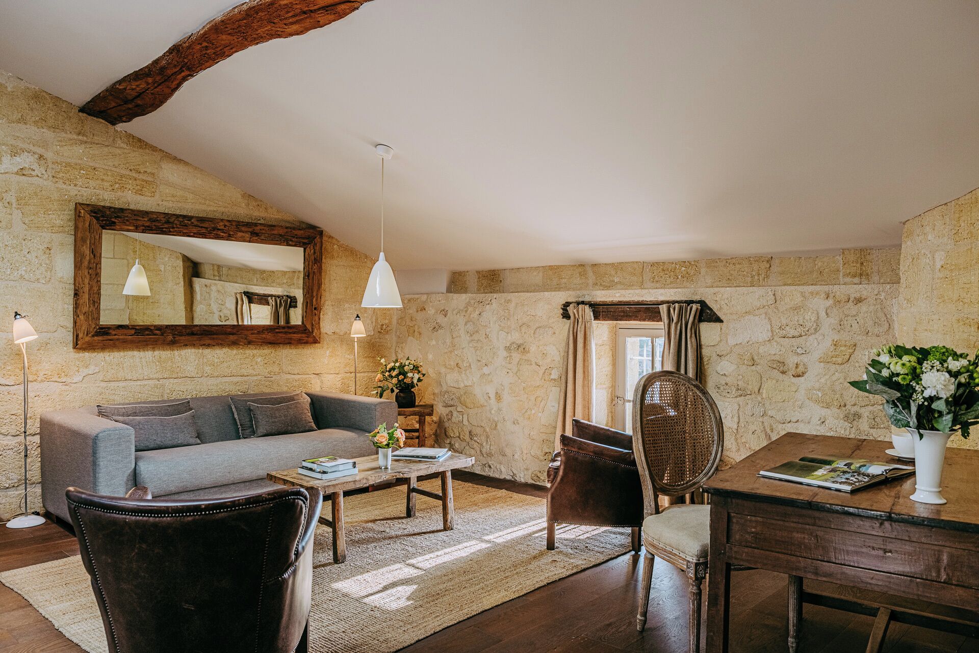 A cozy living room with a stone wall, a wooden ceiling beam, a grey sofa, a coffee table, a wicker chair, and a dining table with a vase of flowers.