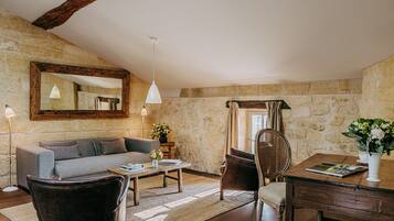 Un salon chaleureux avec un mur en pierre, une poutre en bois au plafond, un canapé gris, une table basse, un fauteuil en osier et une table à manger agrémentée d’un vase de fleurs.