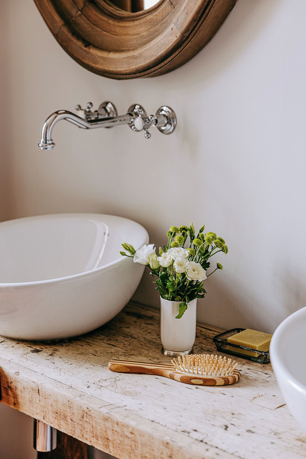 Une salle de bain avec un lavabo blanc, un meuble-lavabo en bois, un robinet en laiton, un vase blanc avec des fleurs, une brosse en bois et un porte-savon.