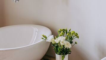 Un baño con un lavamanos blanco, un mueble de tocador de madera, un grifo de latón, un jarrón blanco con flores, un cepillo de madera y un jabonera.