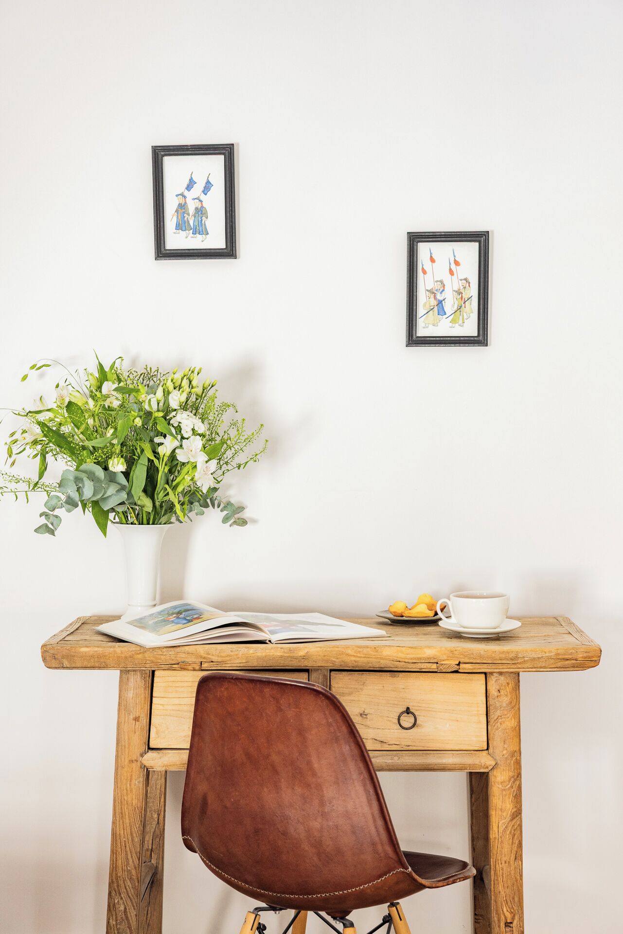 A wooden desk with a vase of flowers, a book, a cup, and a plate of pastries.