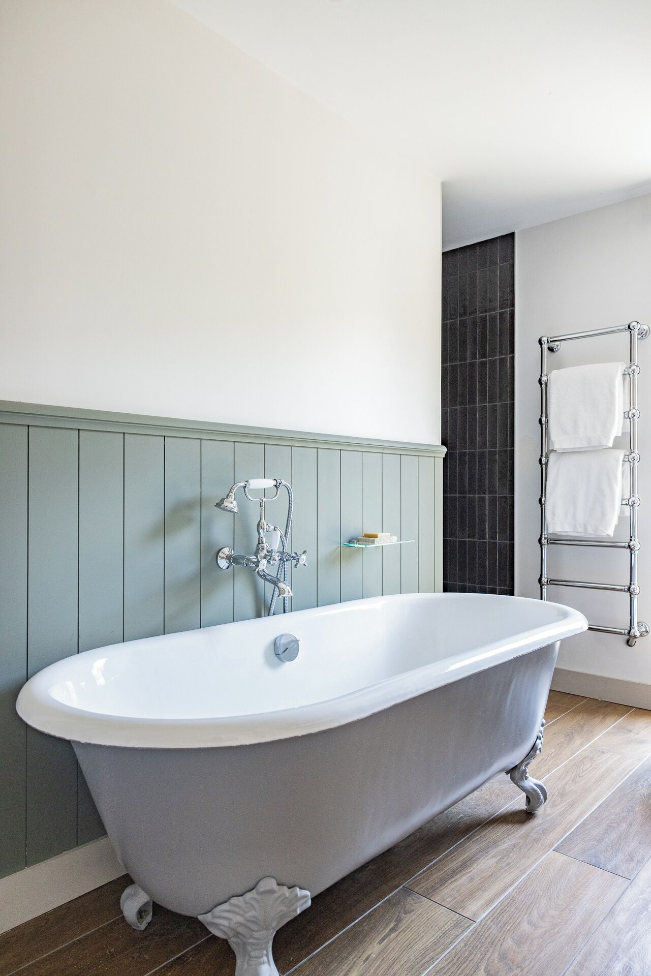 A freestanding clawfoot bathtub with a chrome faucet and shower head, a glass shelf with toiletries, and a towel rack with two towels.