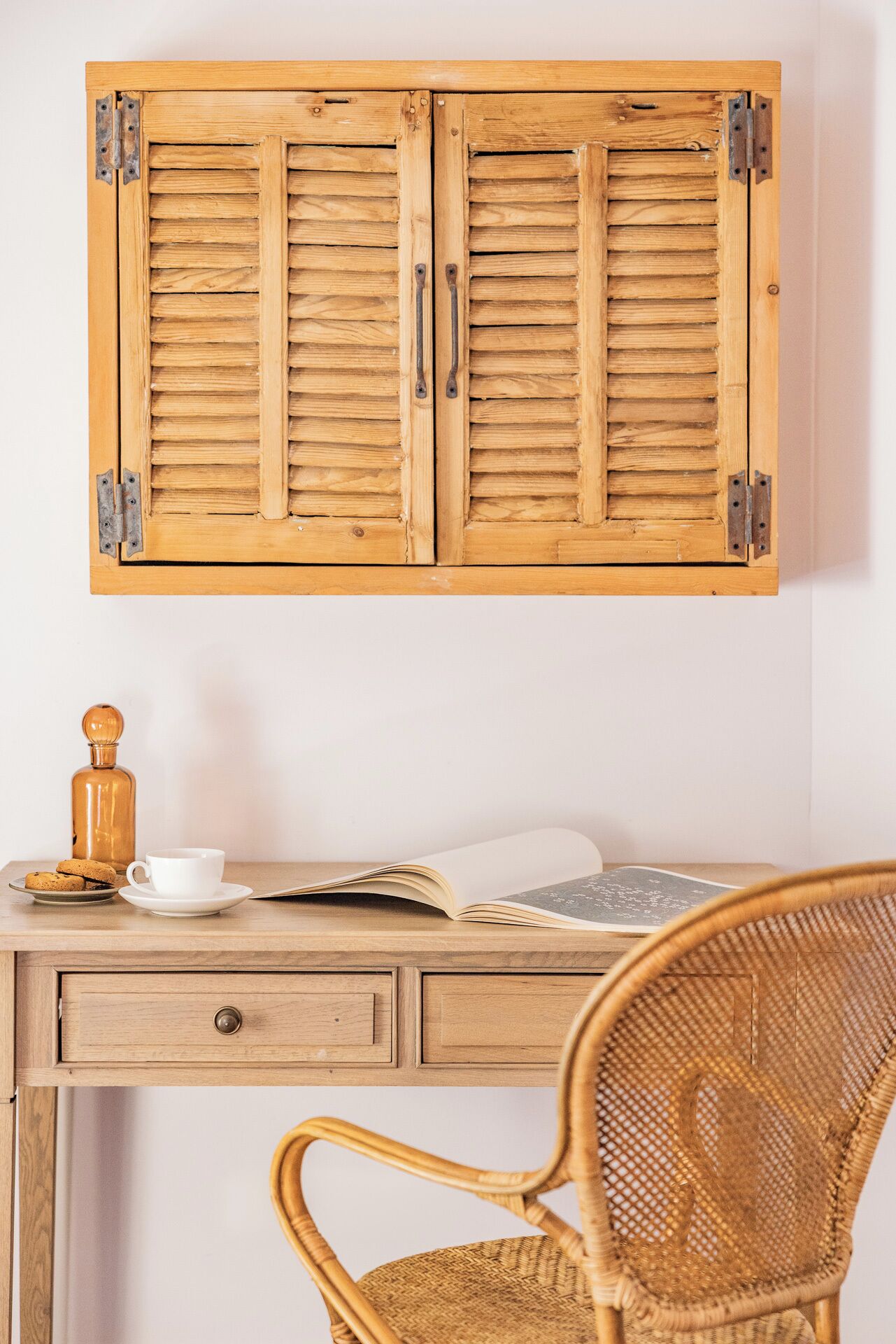 Un mueble de madera con dos puertas, una silla de mimbre, una taza blanca, un libro y una botella sobre una mesa.