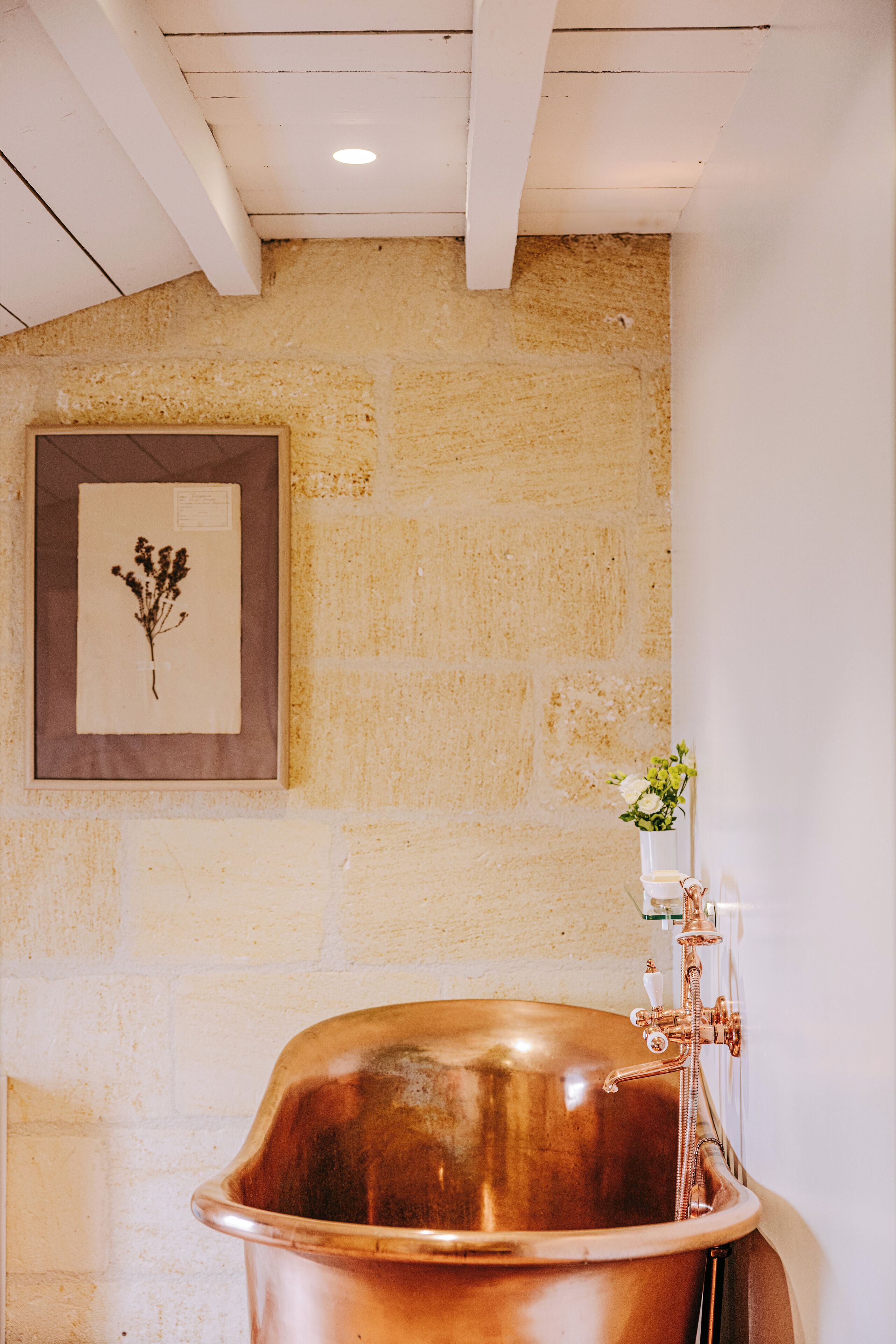 Una bañera de cobre en un baño con pared de piedra e impresión botánica enmarcada.