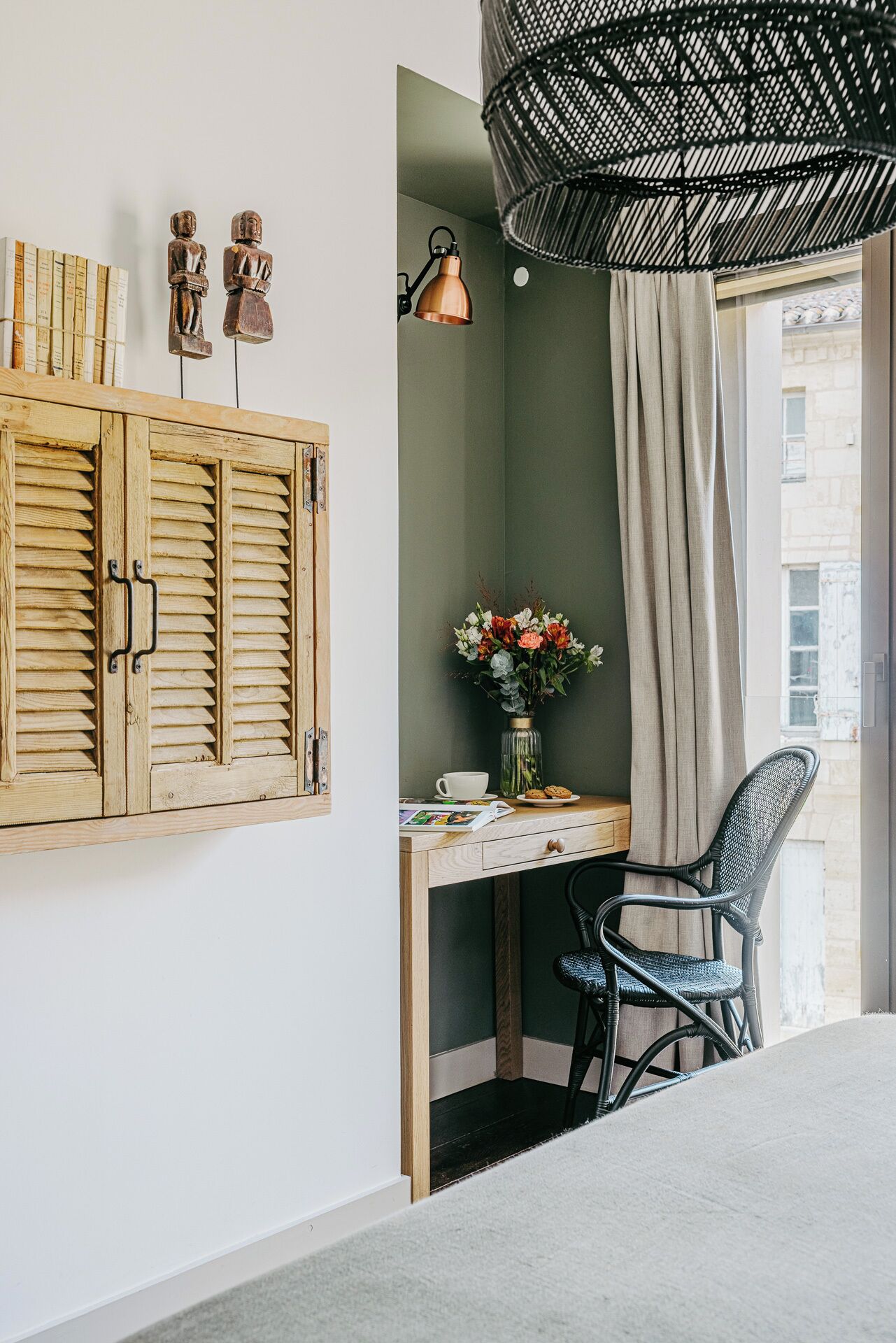 A room with a wooden desk, a wicker chair, a hanging light fixture, and a window with curtains.