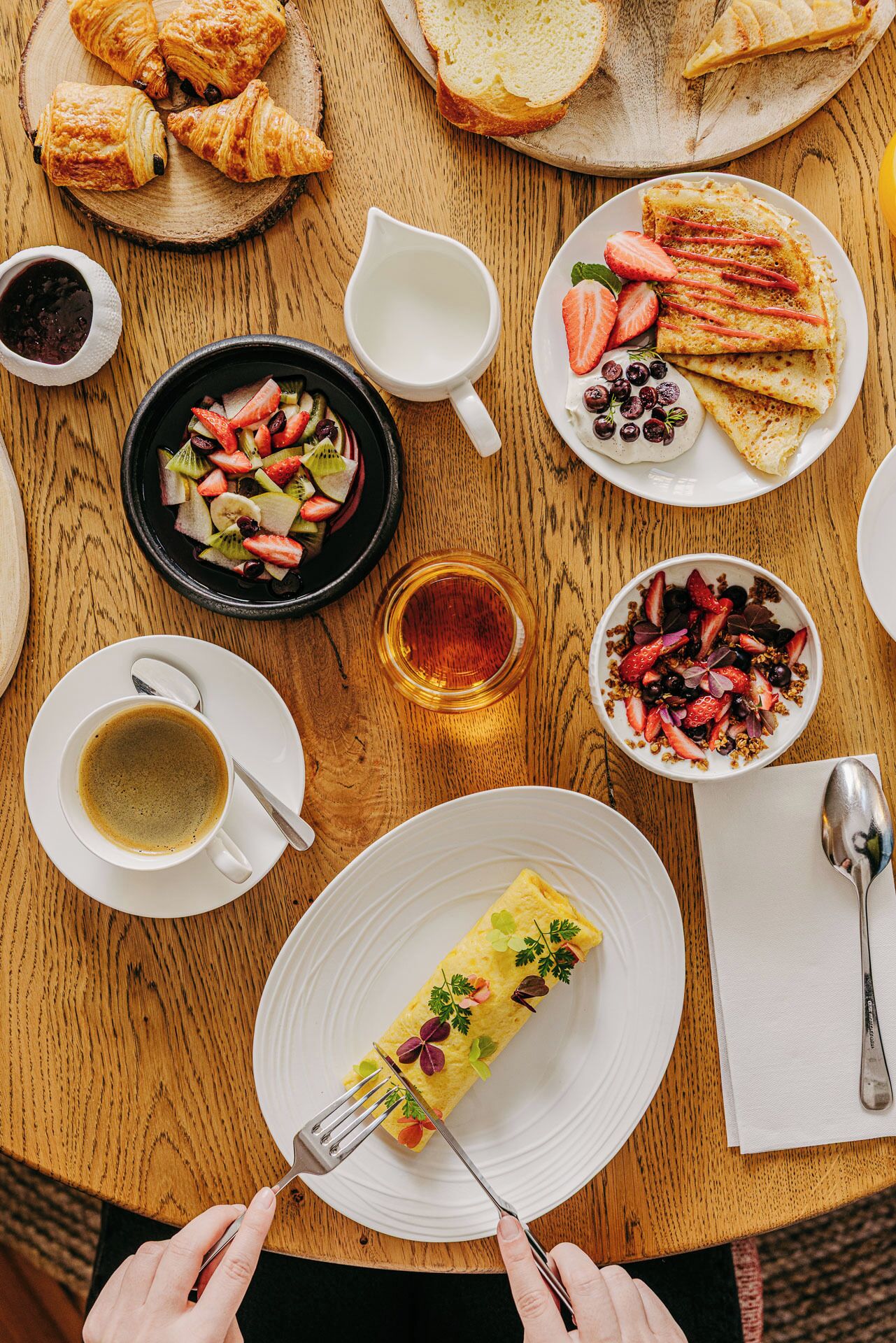 A breakfast setting with croissants, pancakes, a fruit salad, and a rolled omelette on a wooden table.