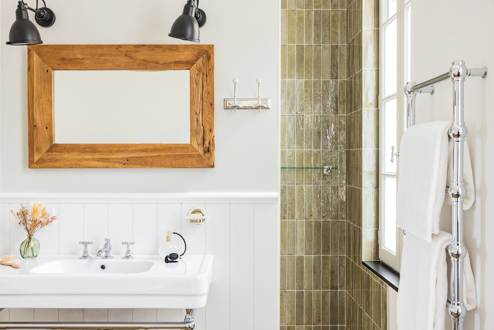 A bathroom with a wooden-framed mirror, a white sink with two faucets, a glass vase with dried flowers, and a towel rack with a white towel.