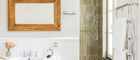 A bathroom with a wooden-framed mirror, a white sink with two faucets, a glass vase with dried flowers, and a towel rack with a white towel.
