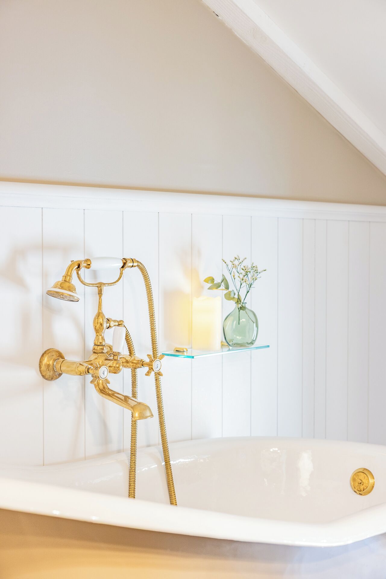Un baño con una bañera blanca, accesorios dorados, un estante de vidrio con una vela y flores, y una pared de azulejos blancos.