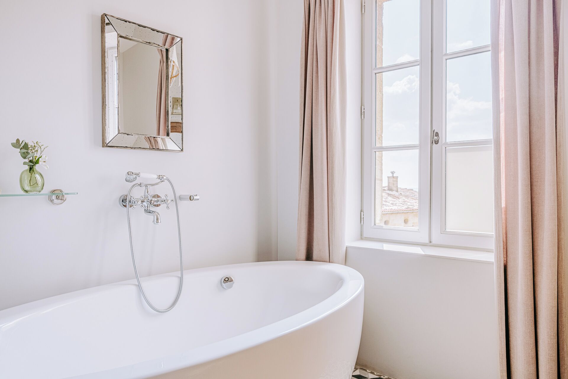A bathroom with a freestanding bathtub, a mirror, a window with curtains, and a small vase with flowers on a shelf.