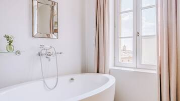 Un baño con una bañera independiente, un espejo, una ventana con cortinas y un pequeño florero con flores en un estante.