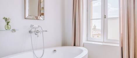 A bathroom with a freestanding bathtub, a mirror, a window with curtains, and a small vase with flowers on a shelf.