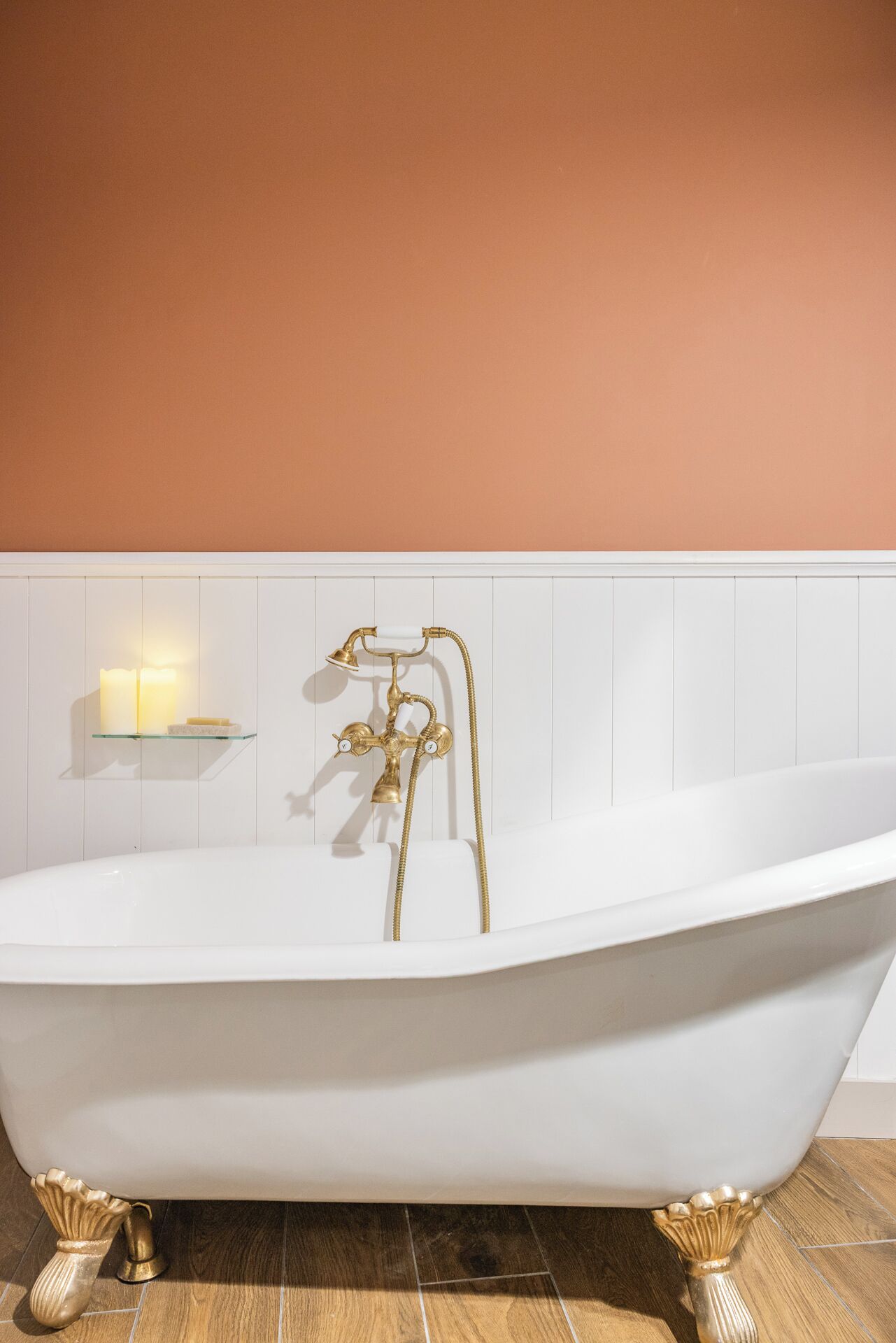 A white freestanding bathtub with gold fixtures, a glass shelf with candles, and a white tiled wall.