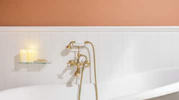A white freestanding bathtub with gold fixtures, a glass shelf with candles, and a white tiled wall.