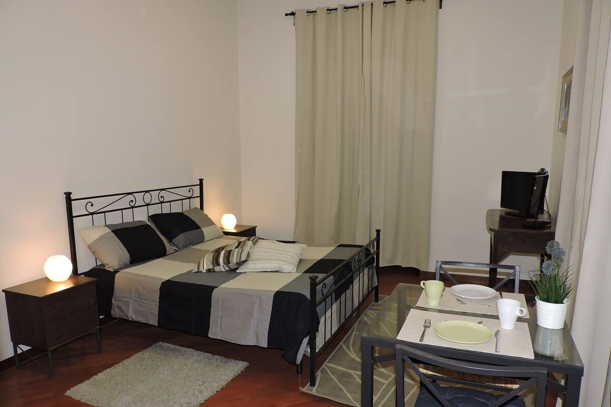 A neatly arranged bedroom with a bed, bedside tables, a small table with plates and cups, and a chair.