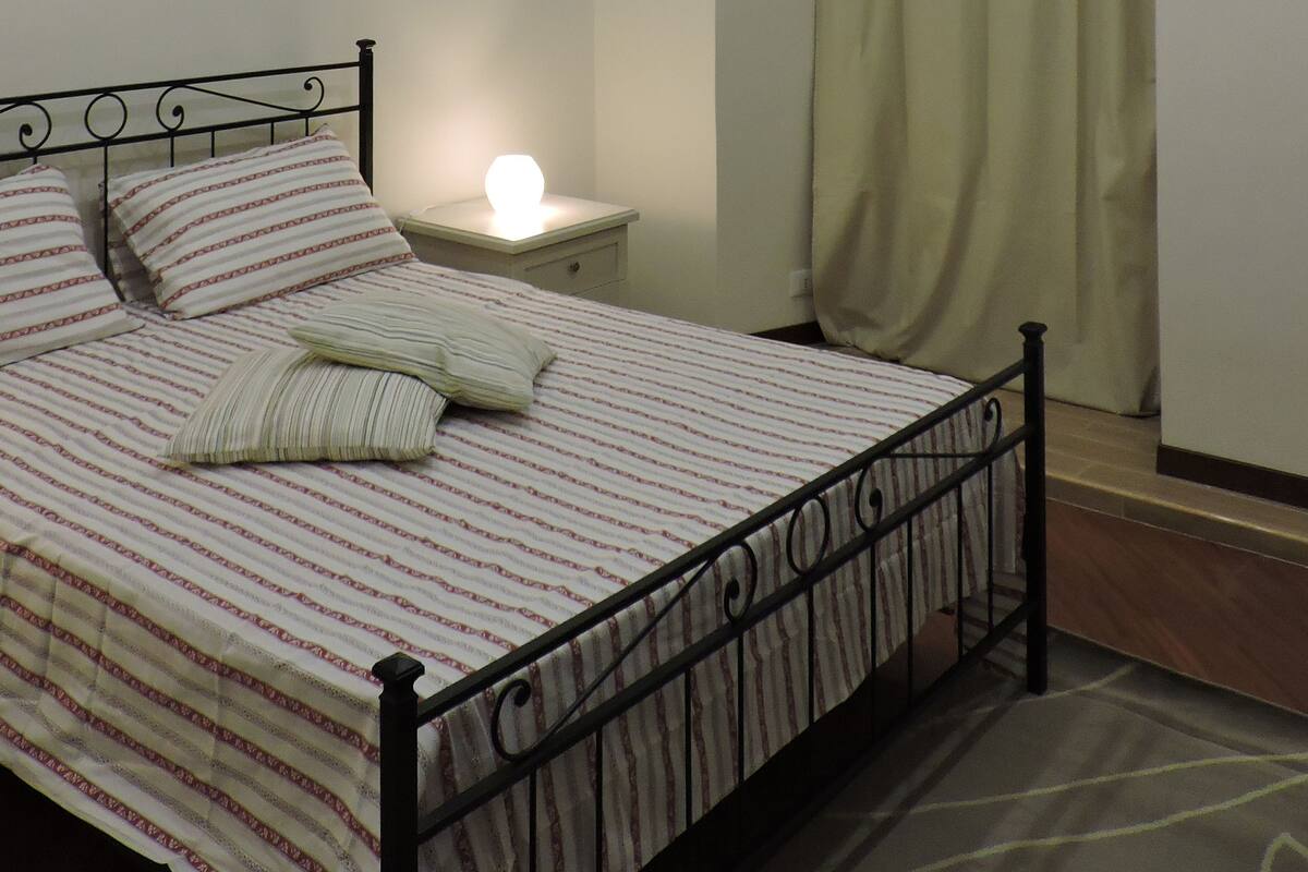 A bedroom with a bed, a nightstand, a lamp, a window with curtains, and a painting on the wall.