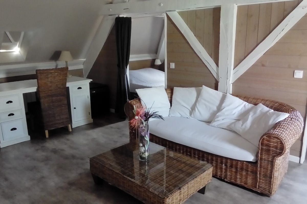 A room with a wicker sofa, a glass coffee table with a decorative item, a white bedside table, a small white cabinet, and a wooden partition wall.