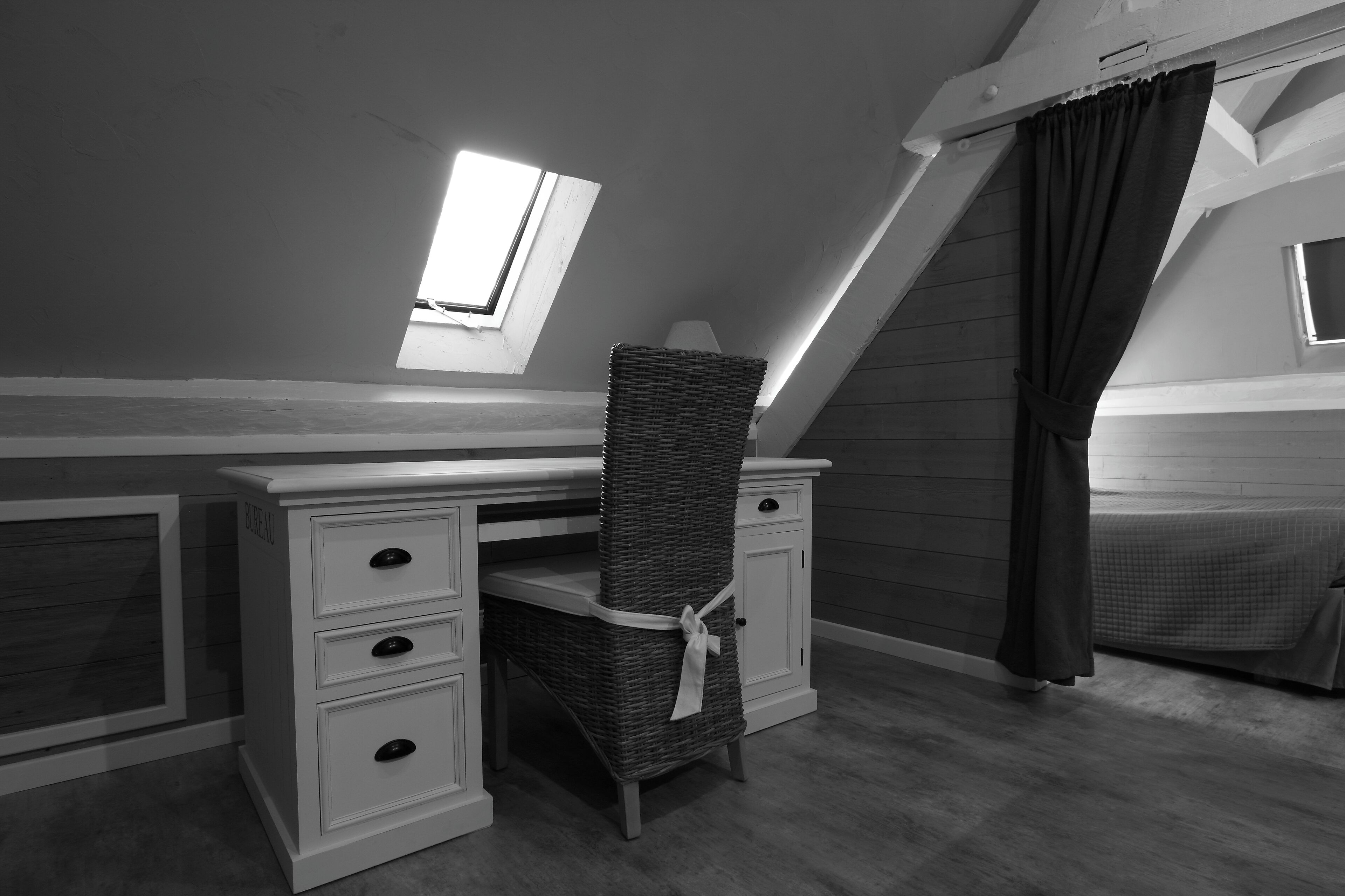 Ein Schlafzimmer mit schrägen Wänden, einem Schreibtisch mit Schubladen, einem Korbstuhl, einem Bett mit kariertem Überwurf und einem Fenster mit Vorhängen.