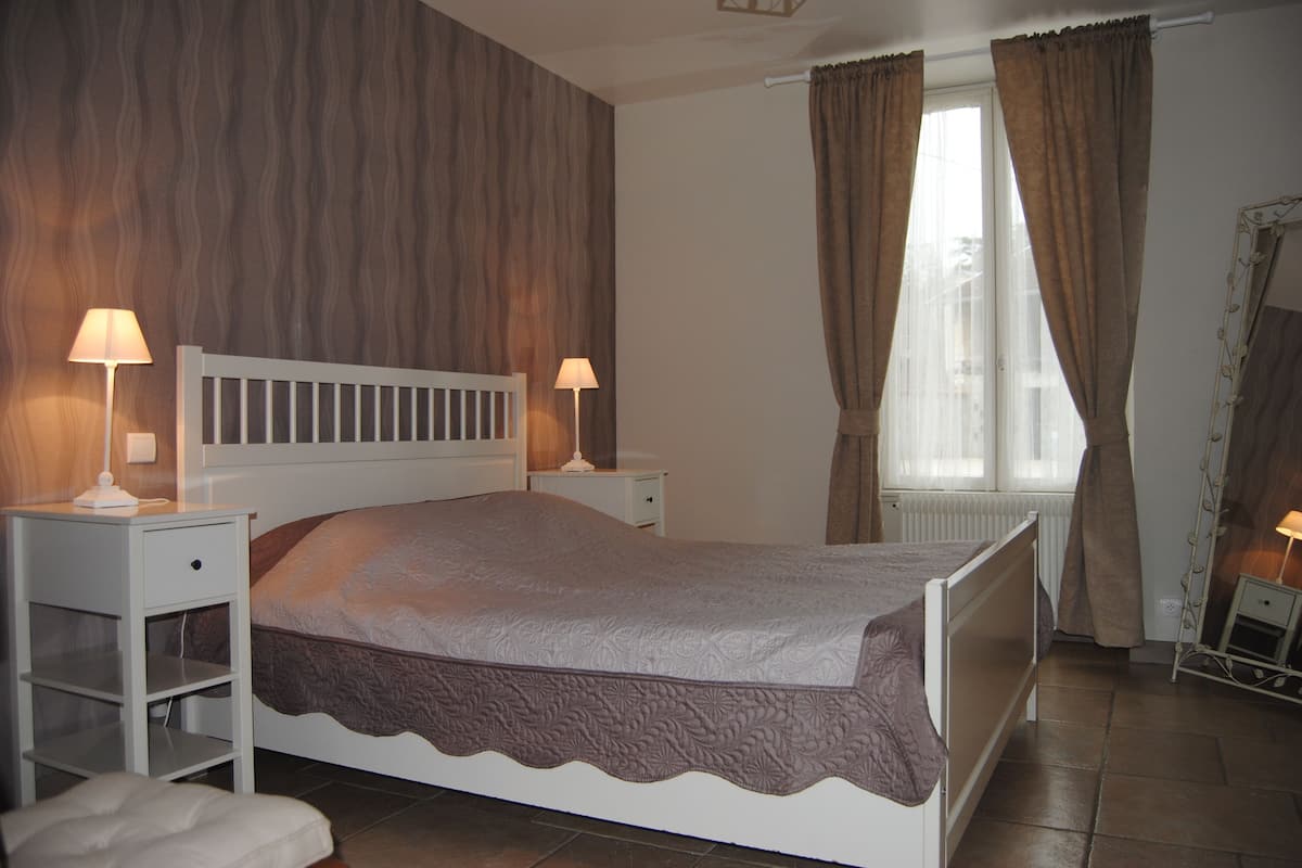A bedroom with a single bed, two bedside tables with lamps, a radiator, a window with curtains, and a mirror on the wall.