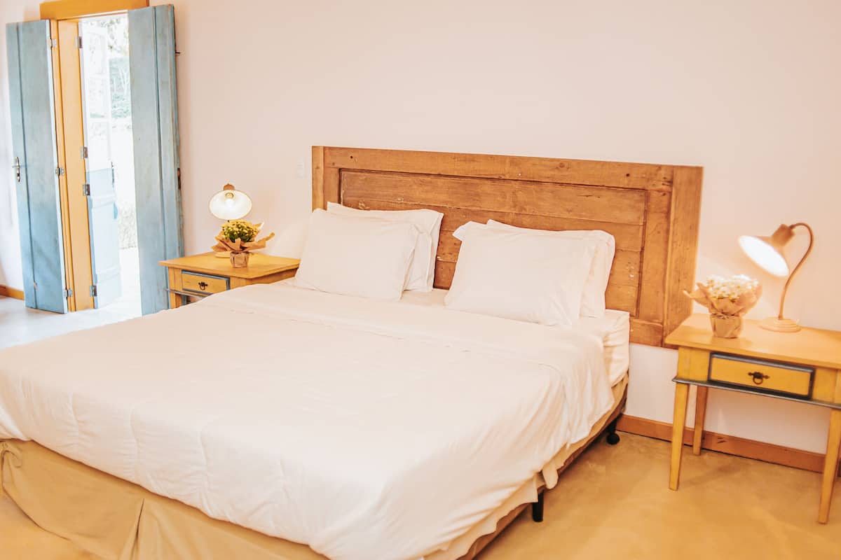 Quarto com cabeceira de madeira, roupa de cama branca, mesa de cabeceira com abajur e criado-mudo com abajur.