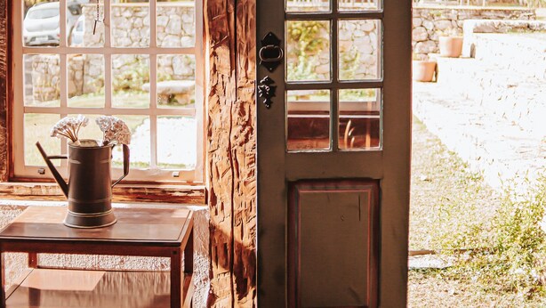 Una puerta de madera con un ventanal de celosías, una jarra metálica sobre una mesa de madera, y una pared de piedra visible a través de los ventanales.