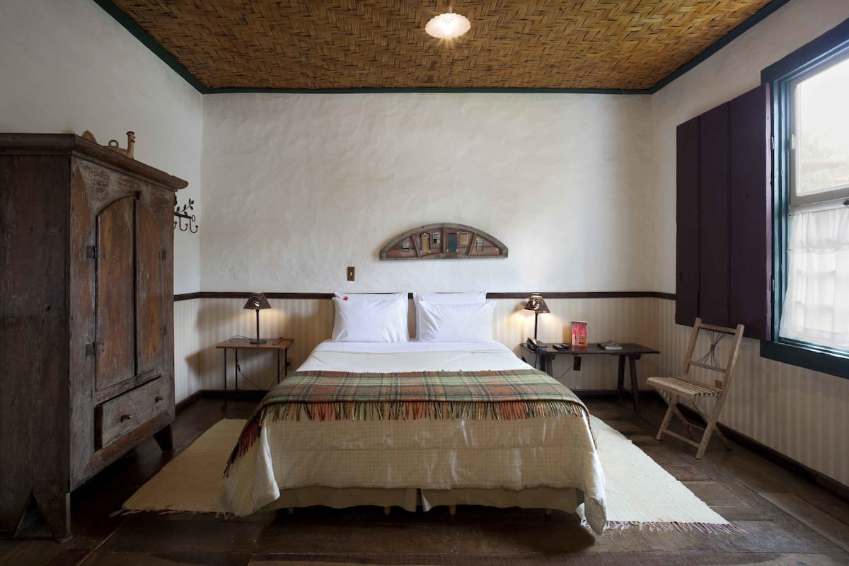 Um quarto com cabeceira de madeira, cama com colcha xadrez, mesa de cabeceira com abajur, armário de madeira e janela com cortinas.