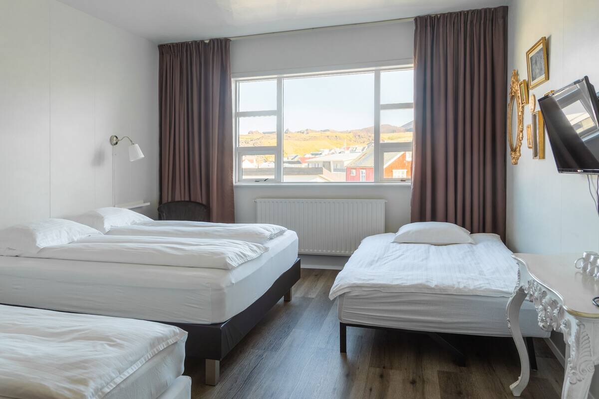 Une chambre d’hôtel avec deux lits simples, un bureau, une chaise, une fenêtre donnant sur des immeubles et un radiateur.