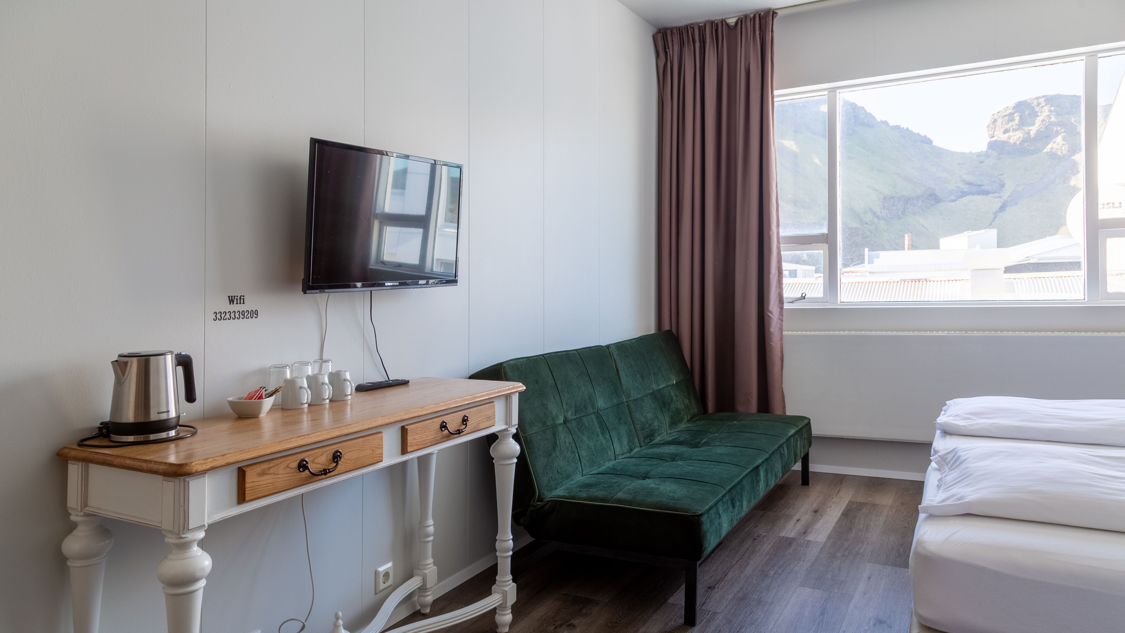 A hotel room with a bed, a green sofa, a wooden desk with a kettle, mugs, and a remote control, a TV mounted on the wall, and a window with a view of mountains.