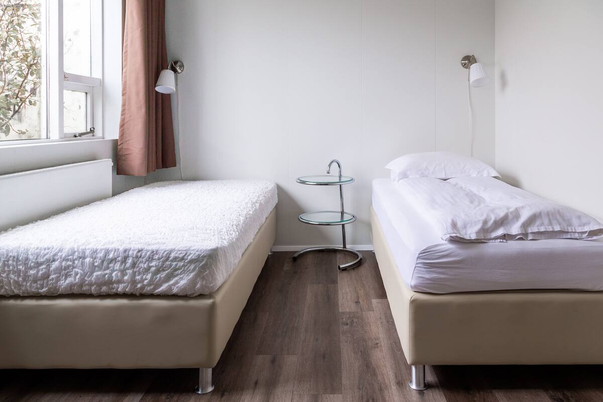 Une chambre avec deux lits simples, une petite table en verre et une fenêtre avec des rideaux.