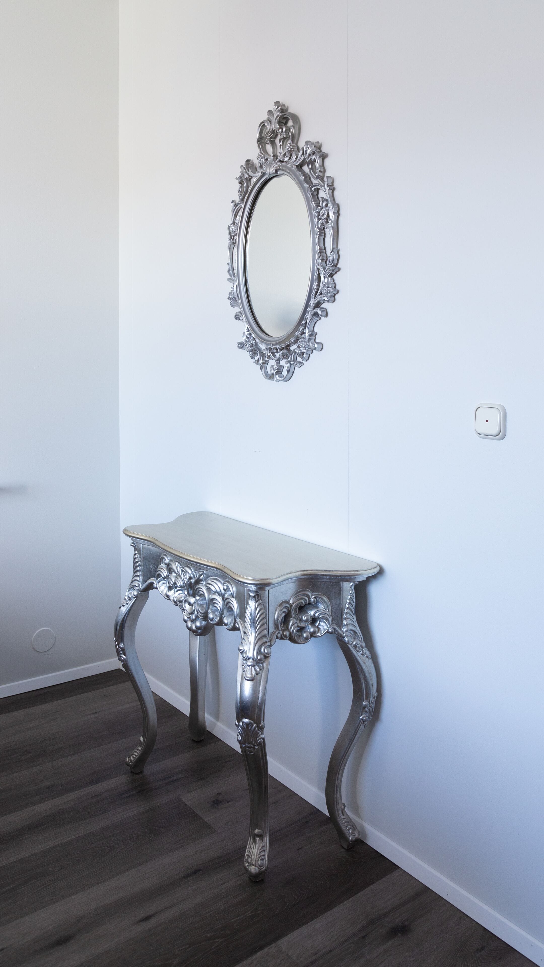 Una habitación con pared blanca, un espejo decorativo con marco plateado y una mesa con marco plateado y patas intrincadas.