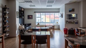 A modern living room with a sofa, a dining table, chairs, a bookshelf, and a large window with a view of buildings. - Guesthouse Hamar (Vestmannaeyjar)