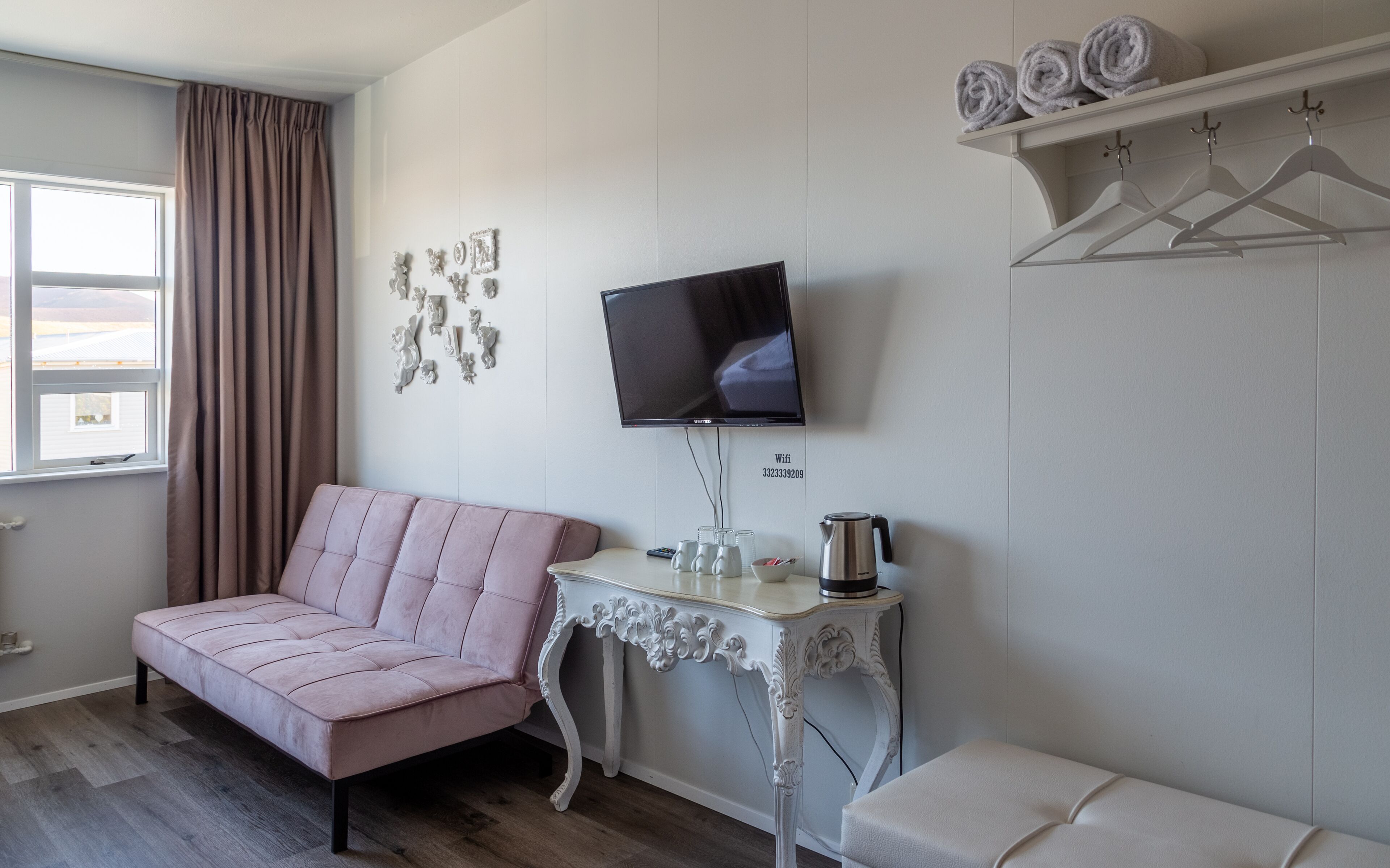 A room with a pink sofa, a television mounted on the wall, a small table with a kettle, and a shelf with towels.