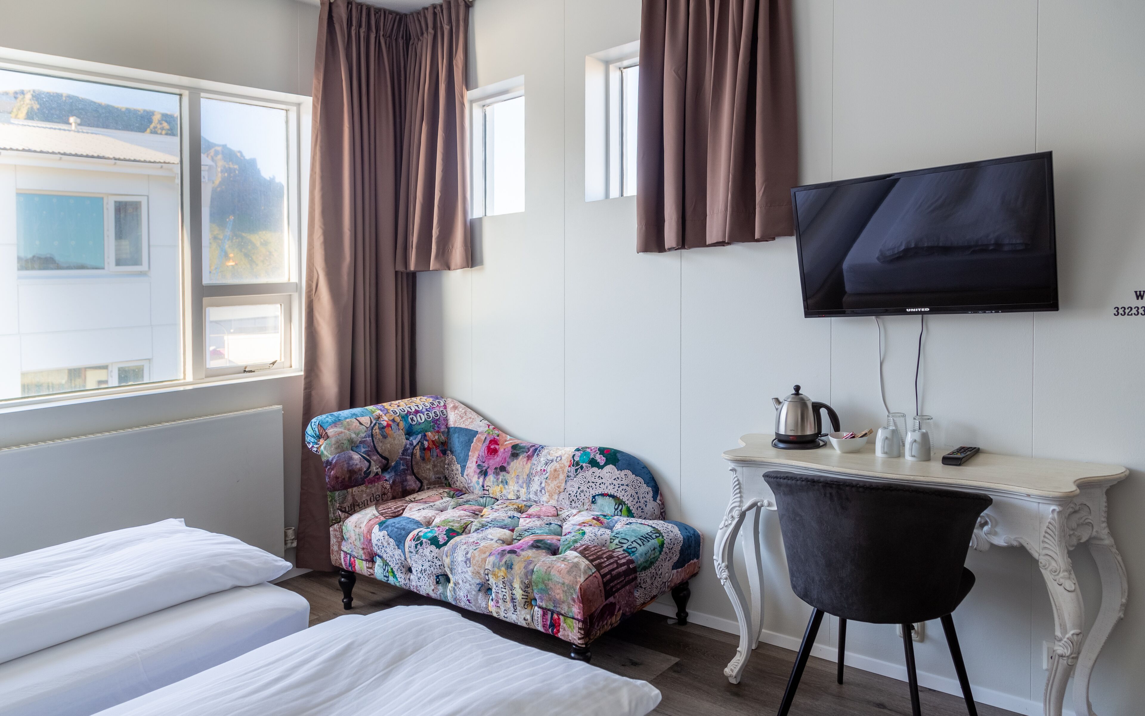 Quarto de hotel com cama, sofá, escrivaninha com chaleira, cadeira e televisão na parede.