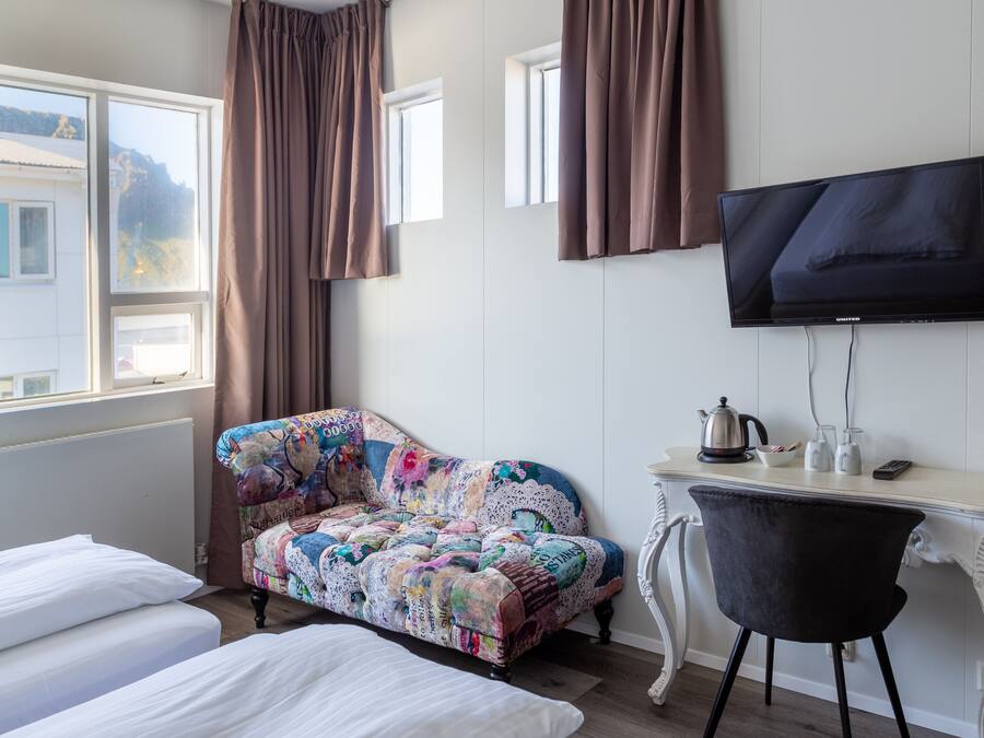 Quarto de hotel com cama, sofá, escrivaninha com chaleira, cadeira e televisão na parede.