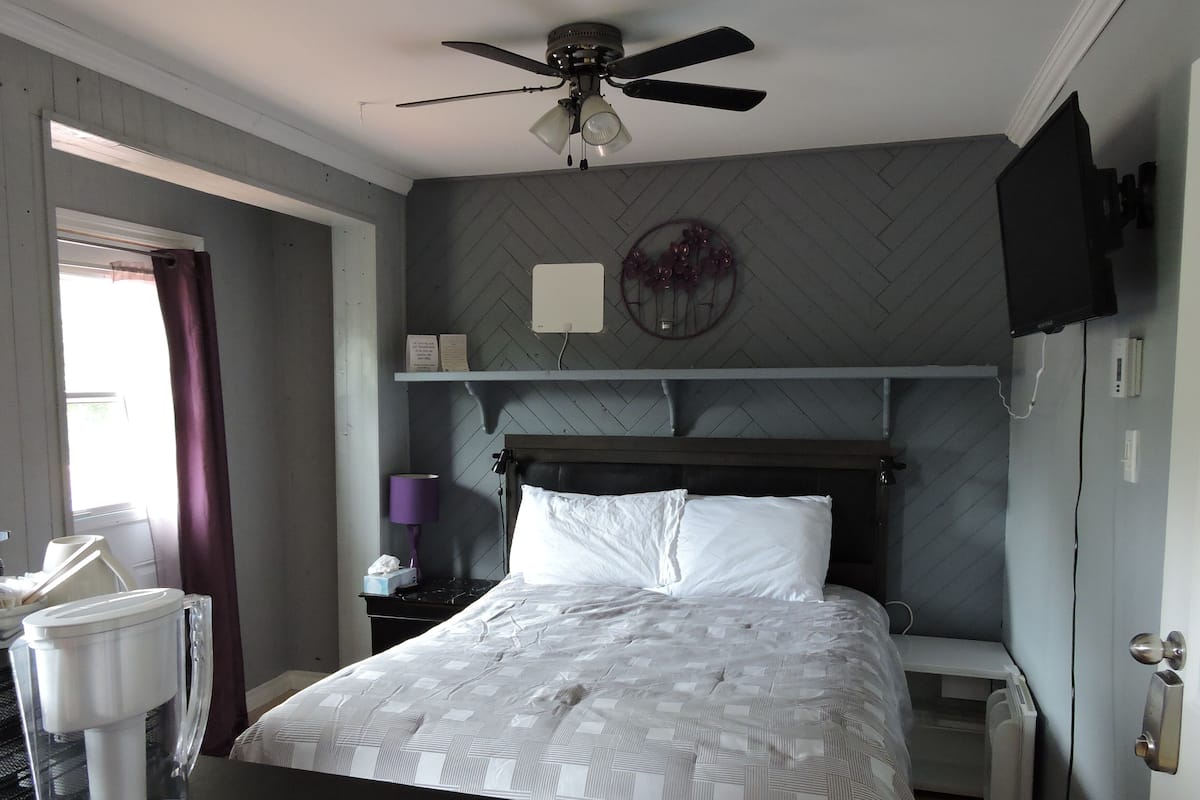 Habitación de hotel con una cama, ventilador de techo, televisor montado, mesita de noche con lámpara y ventana con cortinas.